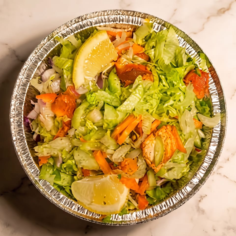 Overhead view of a fresh garden salad with lettuce, tomatoes, cucumbers, carrots, grilled chicken, and lemon wedges in a foil bowl.