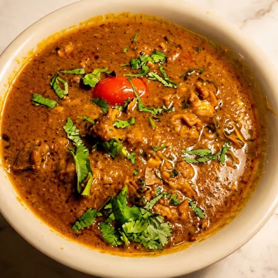 Bowl of rich brown curry garnished with cilantro and cherry tomatoes on a marble tabletop.
